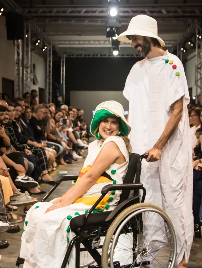 Julia Seixas desfila sorrindo em cadeira de rodas, conduzida na passarela do Friburgo Fashion Week