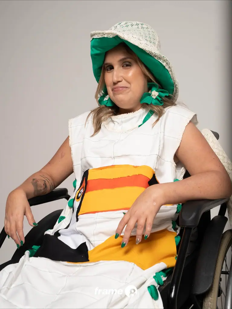 Julia Seixas sorri sentada em sua cadeira de rodas, com chapéu verde e vestido branco do BOOM! Novos Talentos
