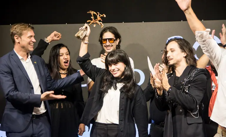 Momento da premiação do BOOM! #01: a vencedora ergue o troféu no palco entre convidados